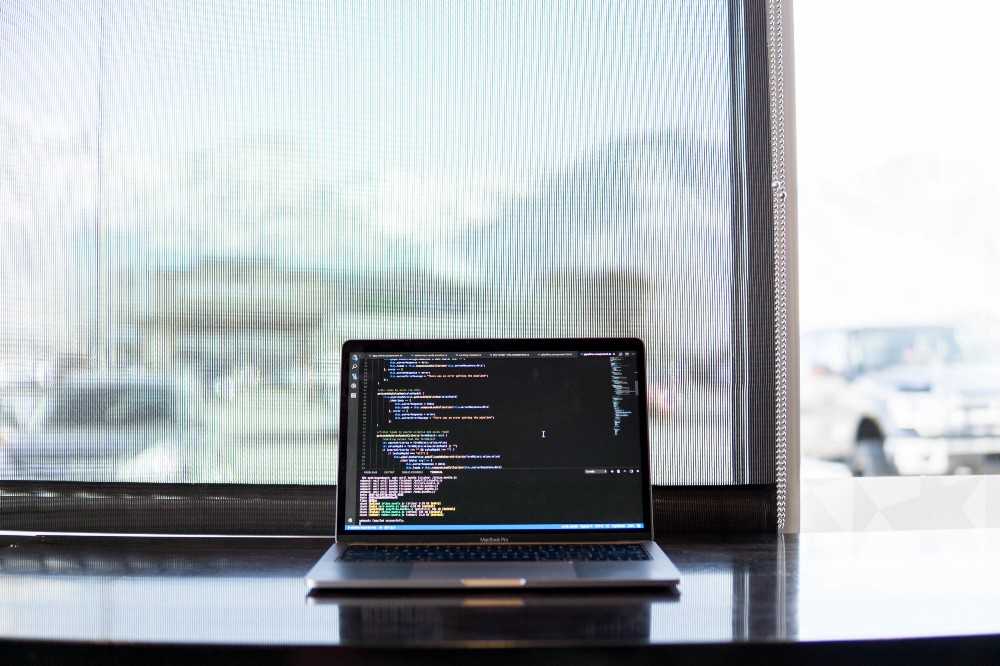 laptop open to code editor on a table in front of a screened window